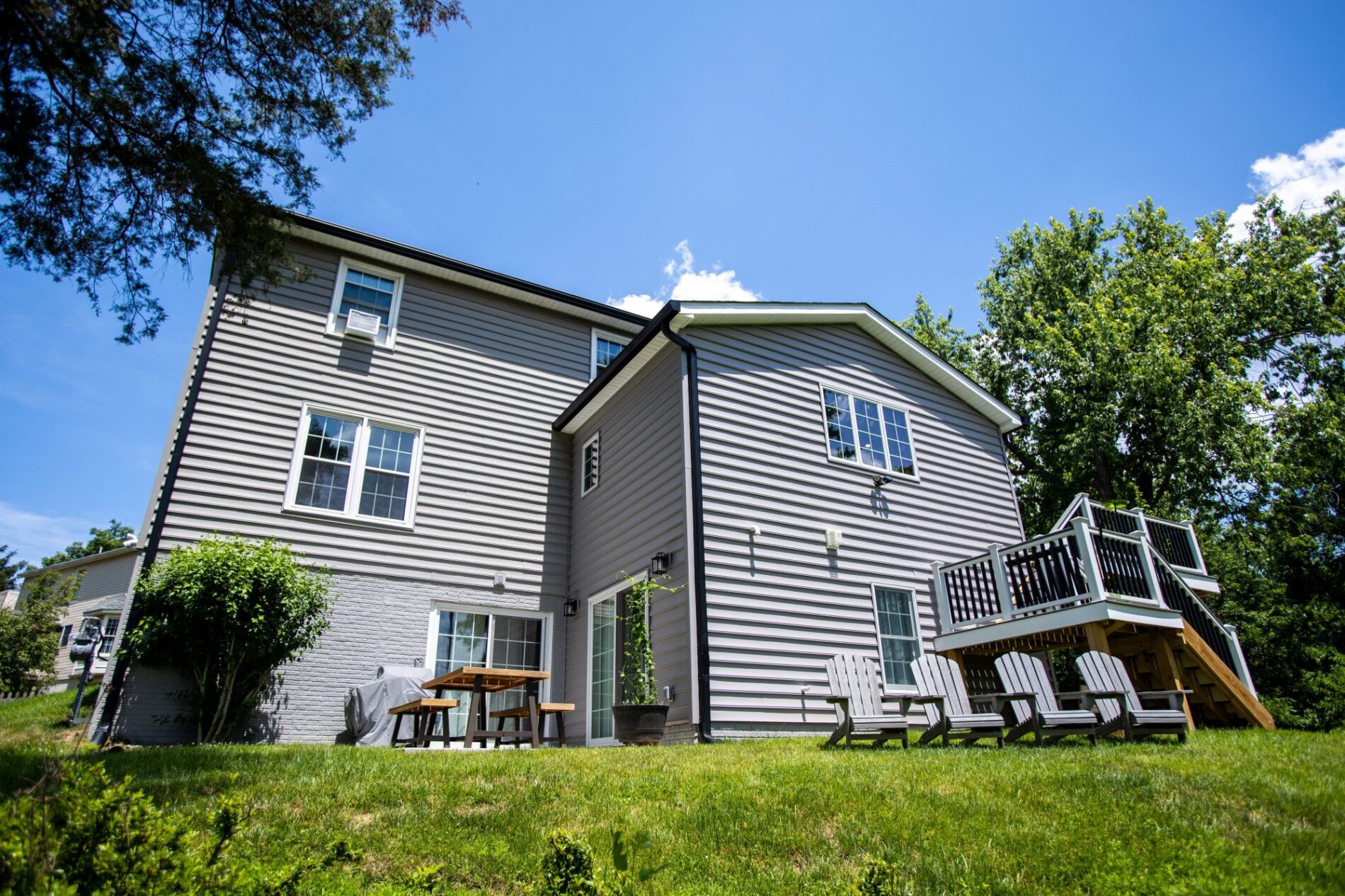 A two-story house with gray siding, multiple windows, and a raised wooden deck with stairs highlights the advantages of design build, featuring Adirondack chairs on a grassy lawn under a clear sky.