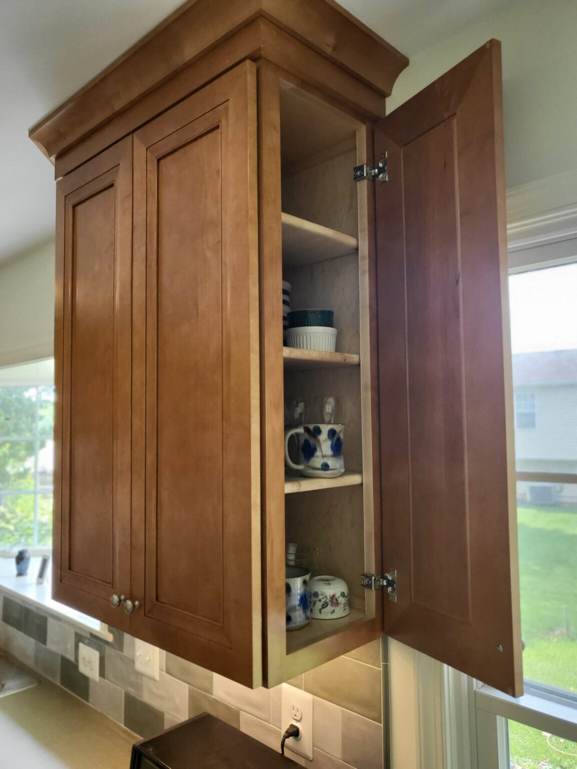 A wooden kitchen cabinet with one door open reveals stacked dishes, mugs, and bowls on three shelves. Sunlight from a window illuminates the scene.