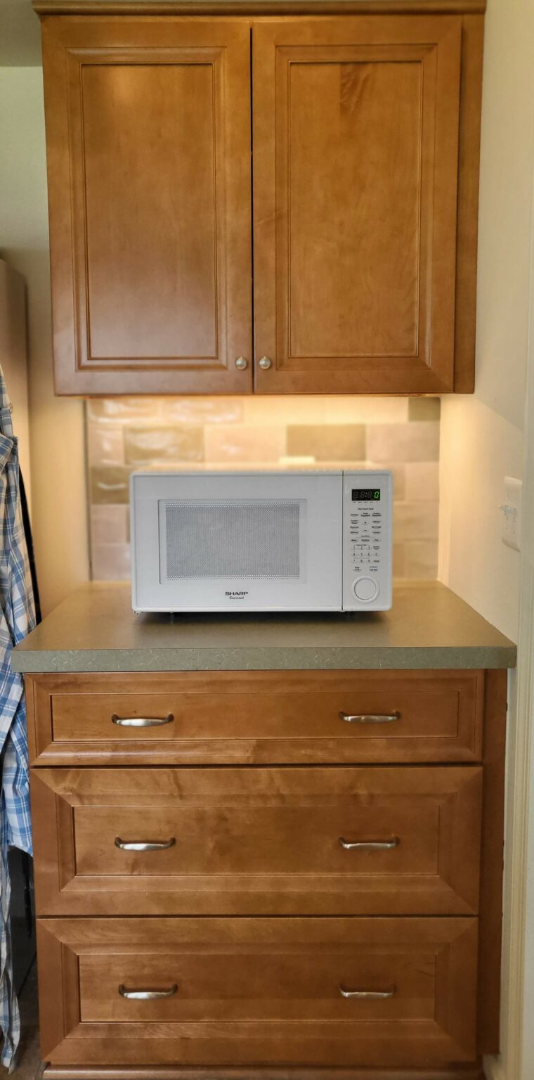 A white microwave is placed on a green countertop between upper and lower wooden cabinets with silver handles in a kitchen.