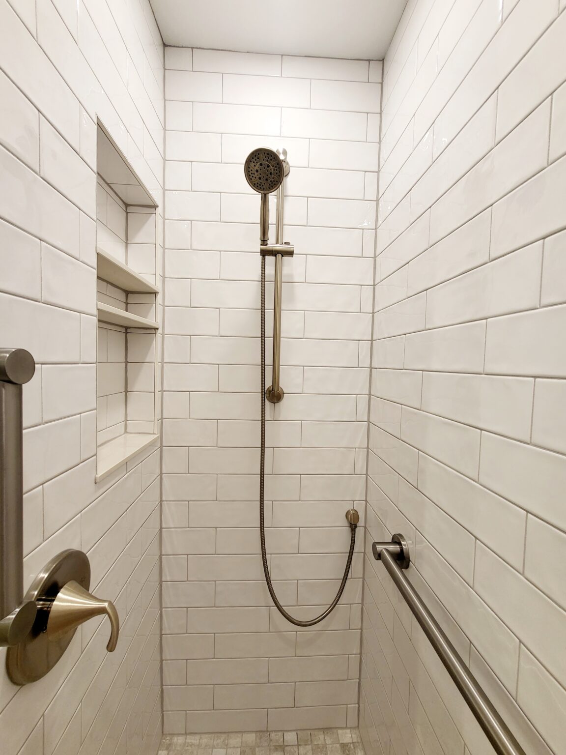 A modern shower with white subway tile walls, a handheld showerhead on a vertical bar, built-in shelves, and metal grab bars.