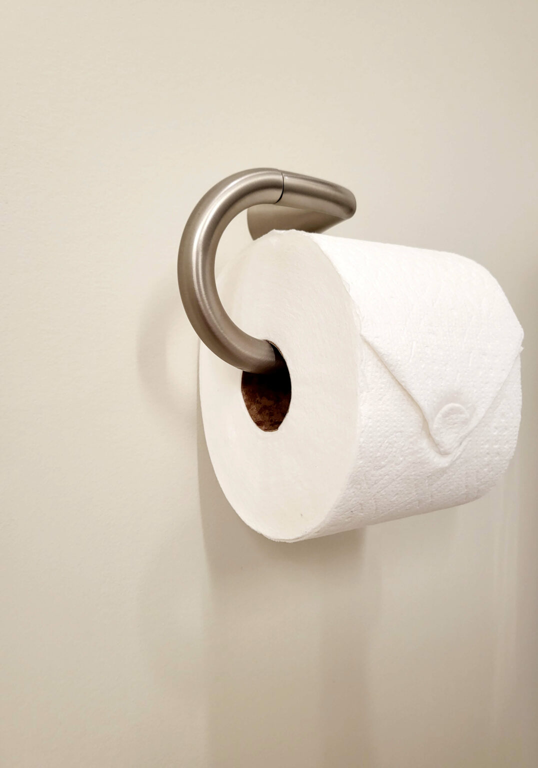 A roll of toilet paper is placed on a silver wall-mounted holder against a plain beige wall.