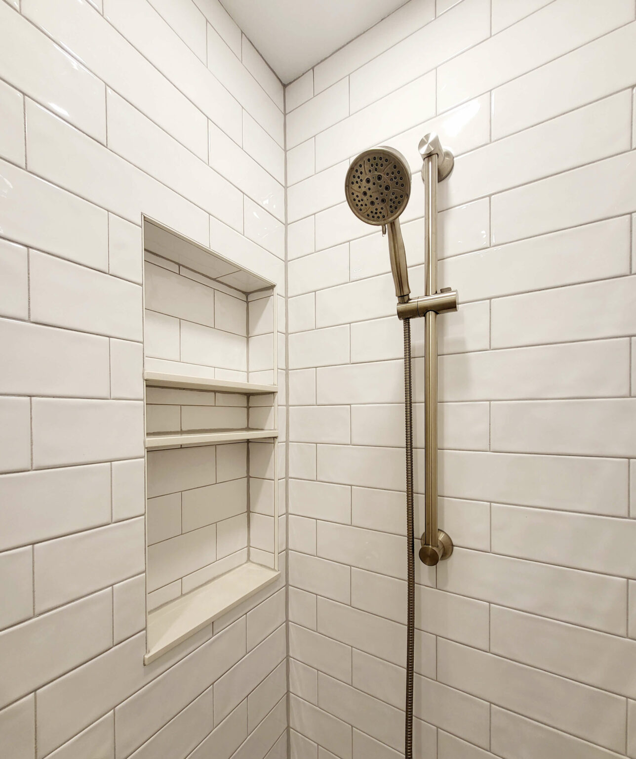 A tiled shower with a built-in niche featuring shelves and a metal adjustable handheld showerhead mounted on a vertical bar.