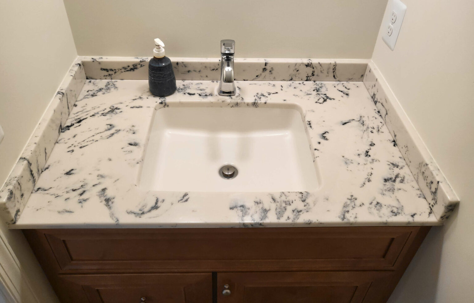 A rectangular bathroom sink with a marbled countertop, chrome faucet, soap dispenser, and wooden vanity cabinet.