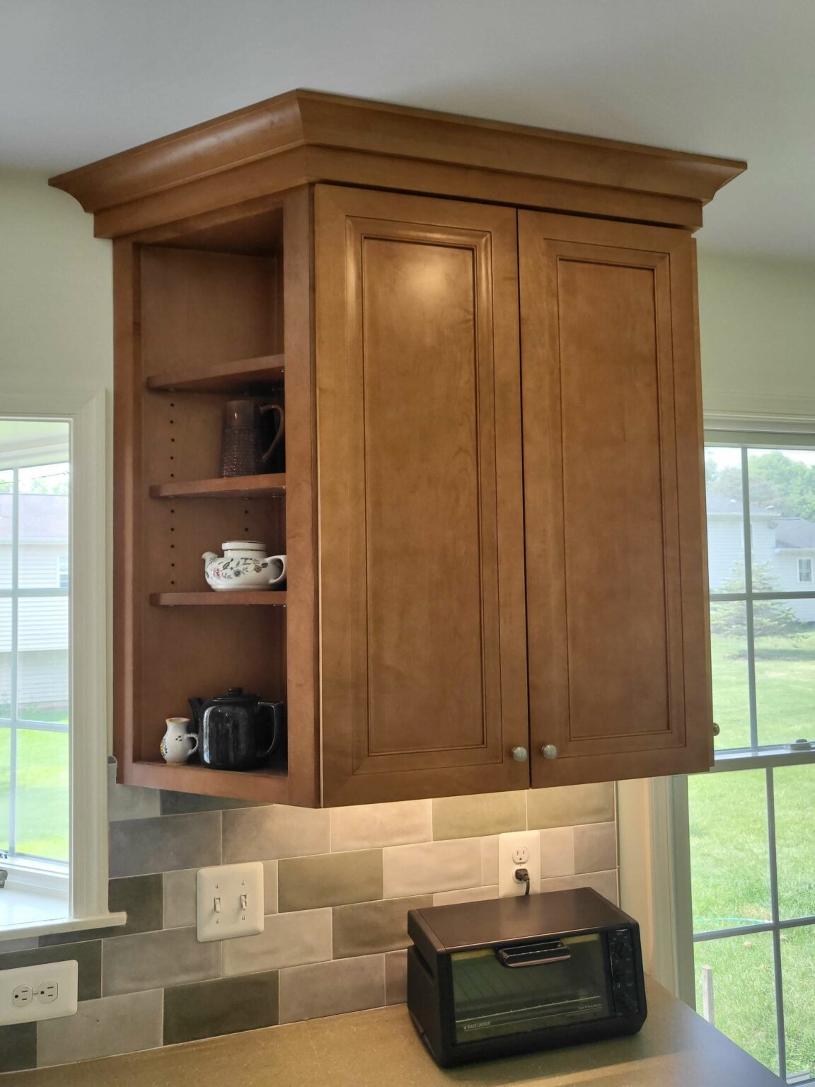 A wooden kitchen cabinet with two closed doors and open shelves on the left side, displaying mugs and a teapot. A toaster oven sits on the counter below.