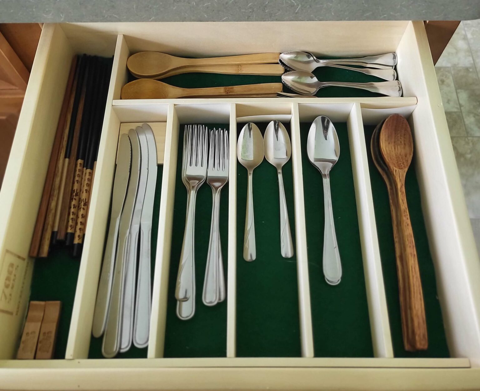 Open kitchen drawer with neatly organized utensils, including knives, forks, spoons, wooden utensils, and chopsticks, arranged in compartments with a green felt lining.