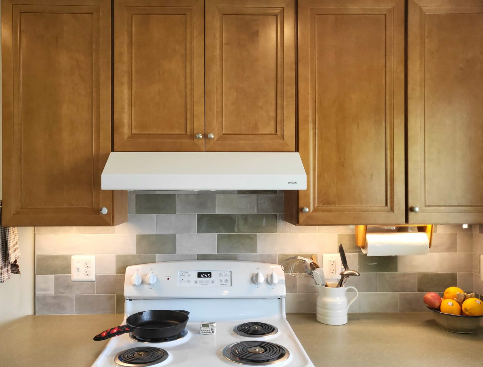 A kitchen stove with a black frying pan, white backsplash, wooden cabinets, a paper towel holder, utensils in a container, and a bowl of fruit on the counter.