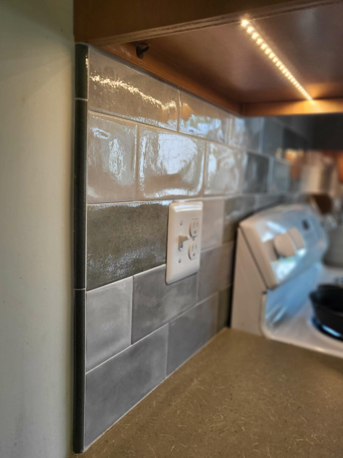 A kitchen backsplash with glossy gray subway tiles, an electrical outlet, a countertop, and part of a stove under a cabinet with LED strip lighting.