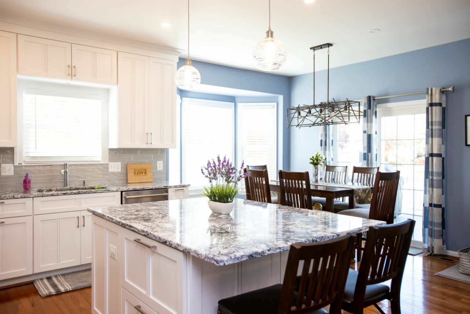 Modern kitchen and dining area with white cabinets, marble countertops, dark wood chairs, pendant lights, and a potted plant on the island. Sunlight enters through large windows.
