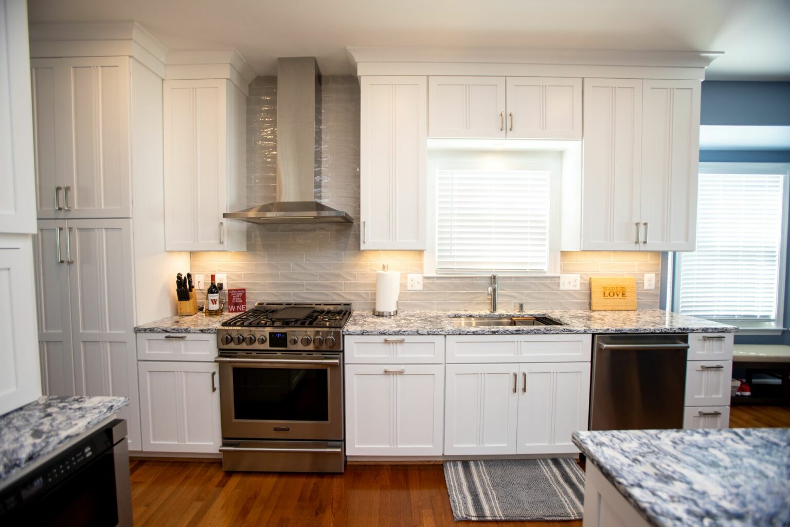 Modern kitchen with white cabinets, stainless steel appliances, marble countertops, a gas stove, sink under a window, and wooden flooring.