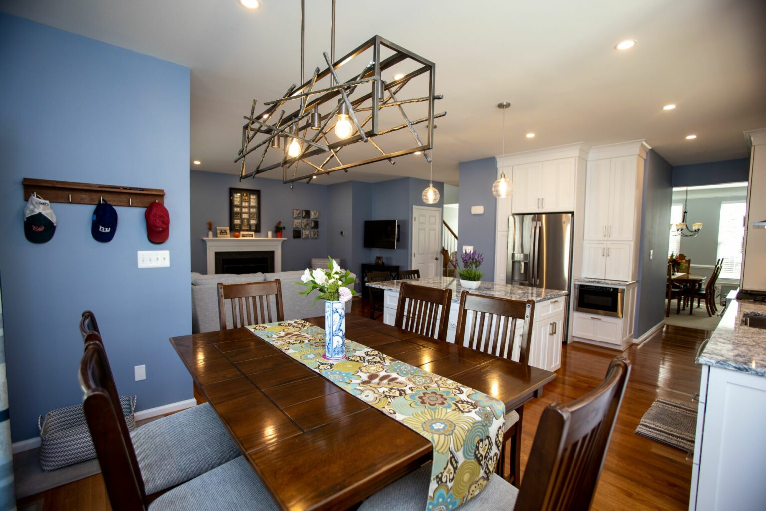 A dining area with a wooden table and chairs, blue walls, a modern light fixture, and an open view of the kitchen and living spaces in a well-lit home.