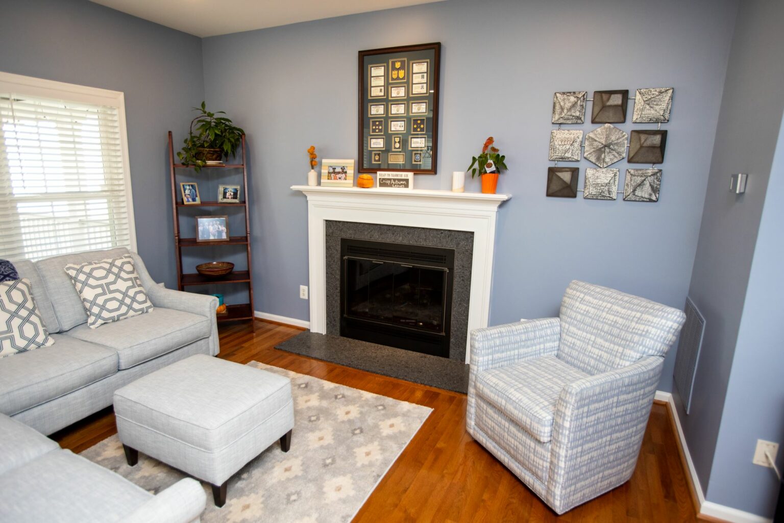 A living room with a gray sofa, matching ottoman, patterned armchair, fireplace with white mantel, blue walls, wall art, and a wooden floor.