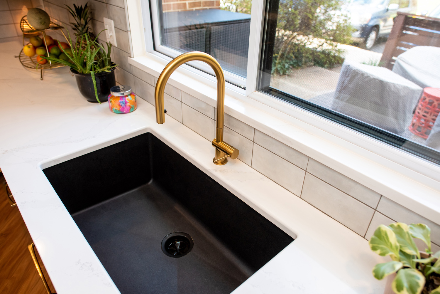 A modern kitchen sink with a black basin, gold faucet, white marble countertop, plants, and a fruit bowl near a window.