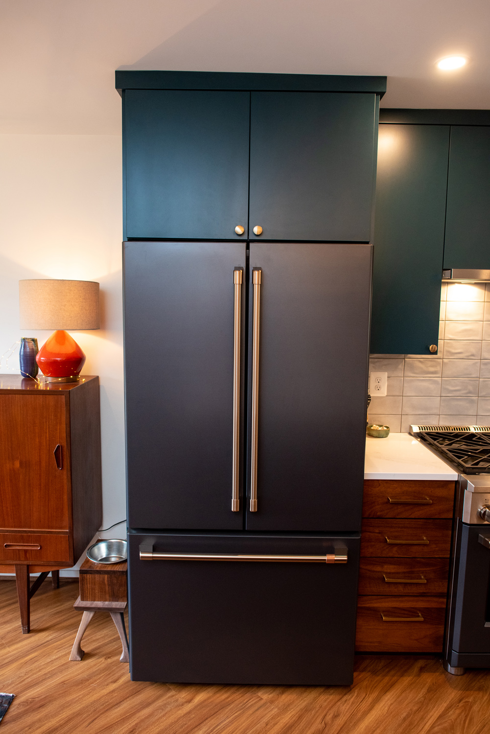 A modern black refrigerator with double doors and a bottom freezer is built into a kitchen with dark cabinets and wooden flooring.
