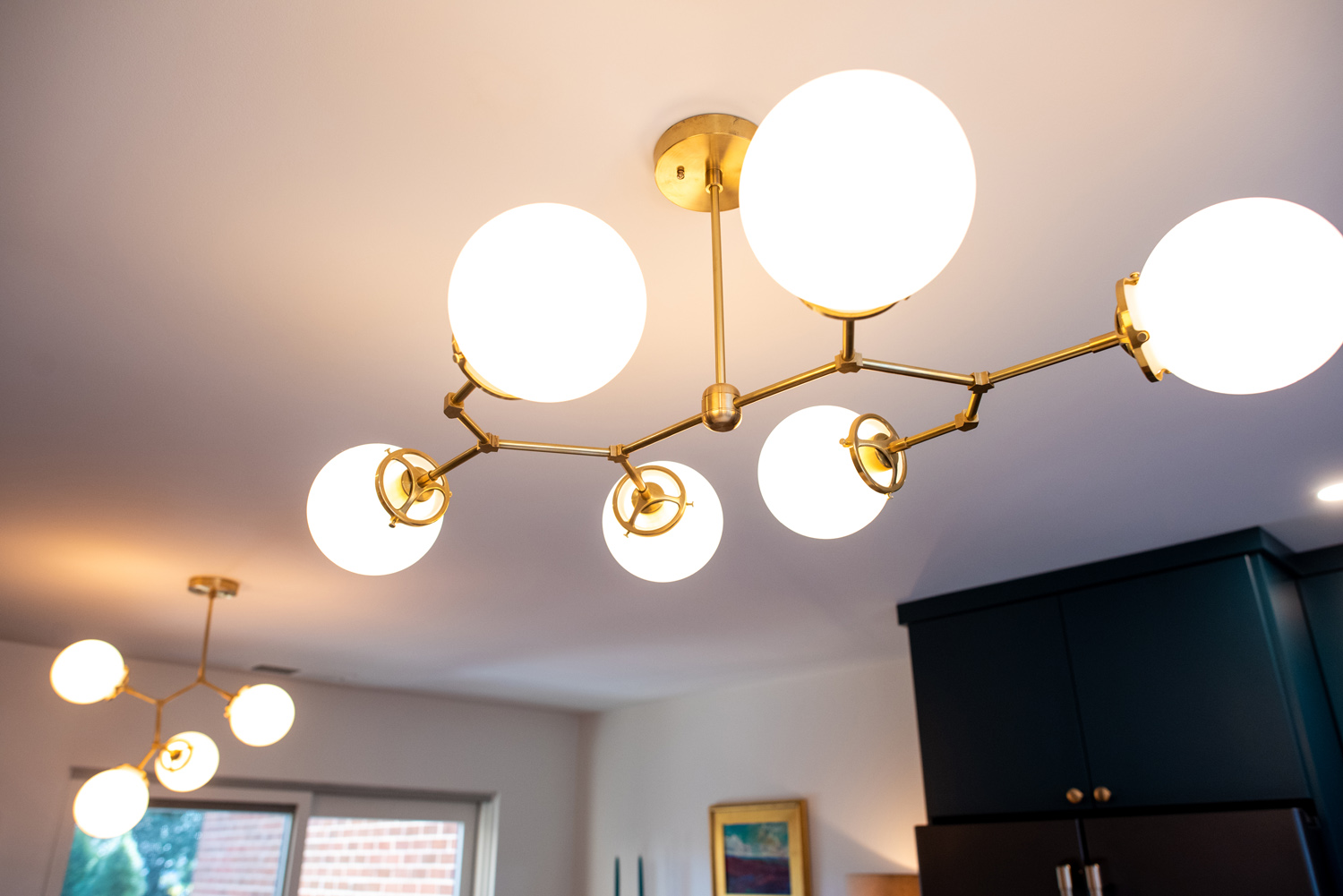 Modern brass chandelier with six round white glass globes is mounted on a ceiling, with a similar smaller fixture visible in the background.