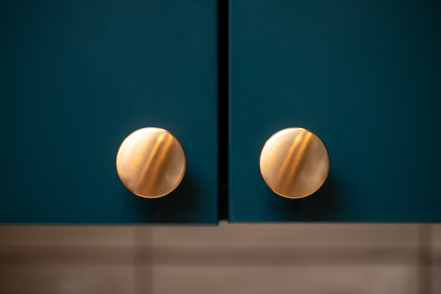 Close-up of two matte gold round cabinet knobs on dark teal cabinet doors.
