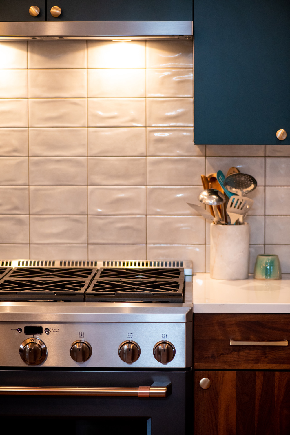 A stainless steel stove with four knobs sits beneath a tiled backsplash. Utensils in a white holder and a turquoise glass are on the counter to the right.