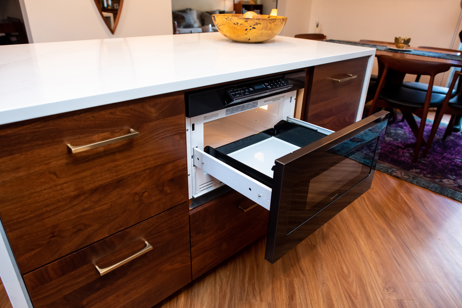 A kitchen island with a built-in microwave drawer open, surrounded by wooden cabinetry and a bowl on the countertop.