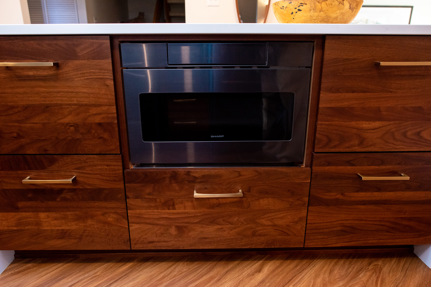 Built-in stainless steel microwave oven set in dark wood kitchen cabinetry with gold handles and a white countertop.