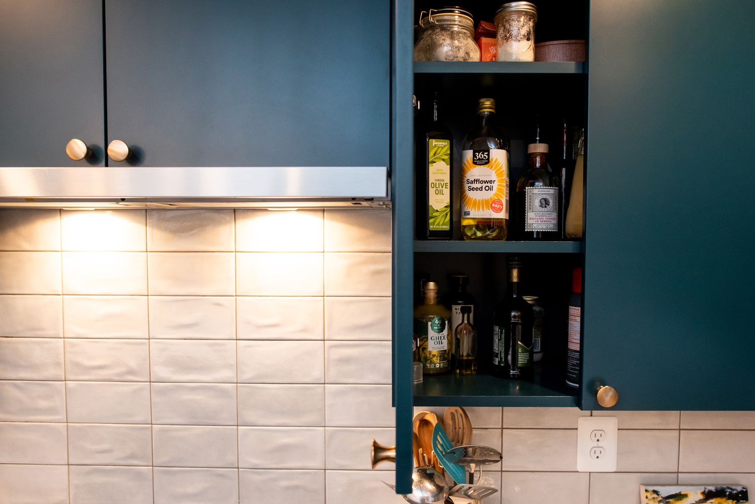 Open kitchen cabinet displaying bottles of cooking oils and seasonings next to a tiled backsplash and blue cupboard doors.