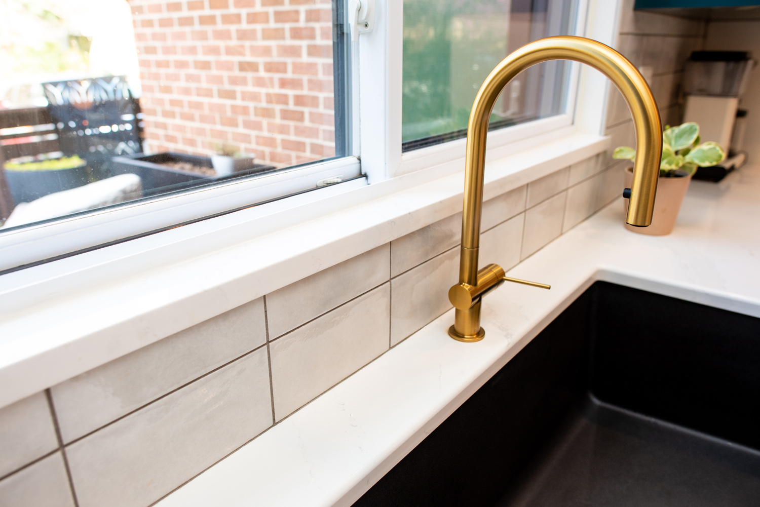 A modern kitchen sink with a gold faucet, white countertop, light gray tiled backsplash, and a small potted plant by the window.