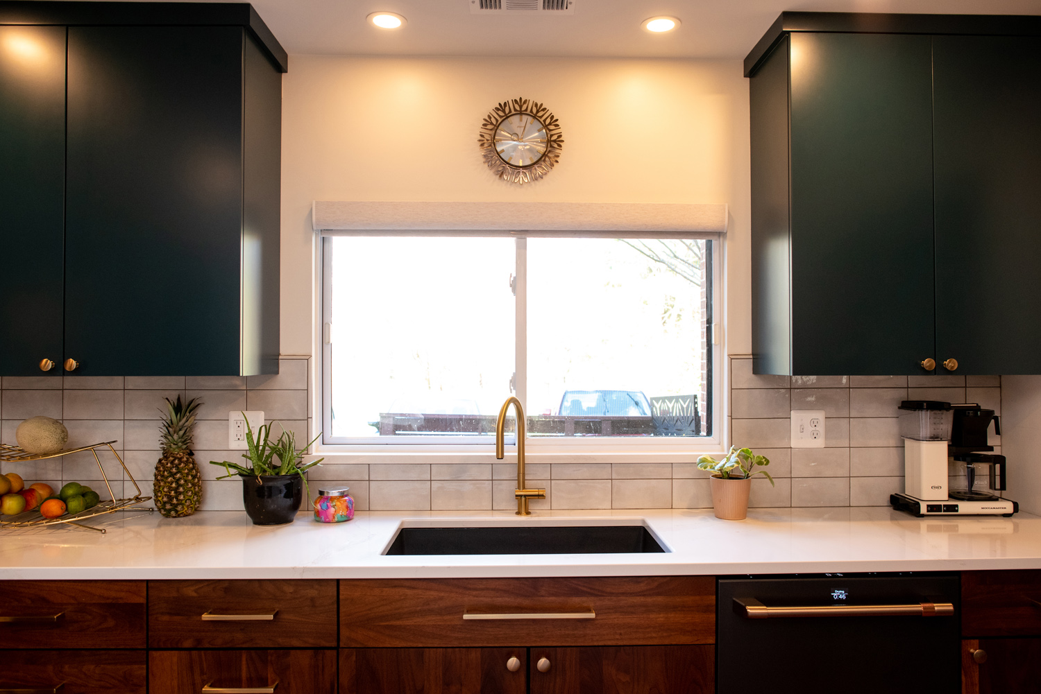 Modern kitchen with dark cabinets, white countertop, gold faucet, fruit basket, potted plants, coffee maker, and window above the sink letting in natural light.
