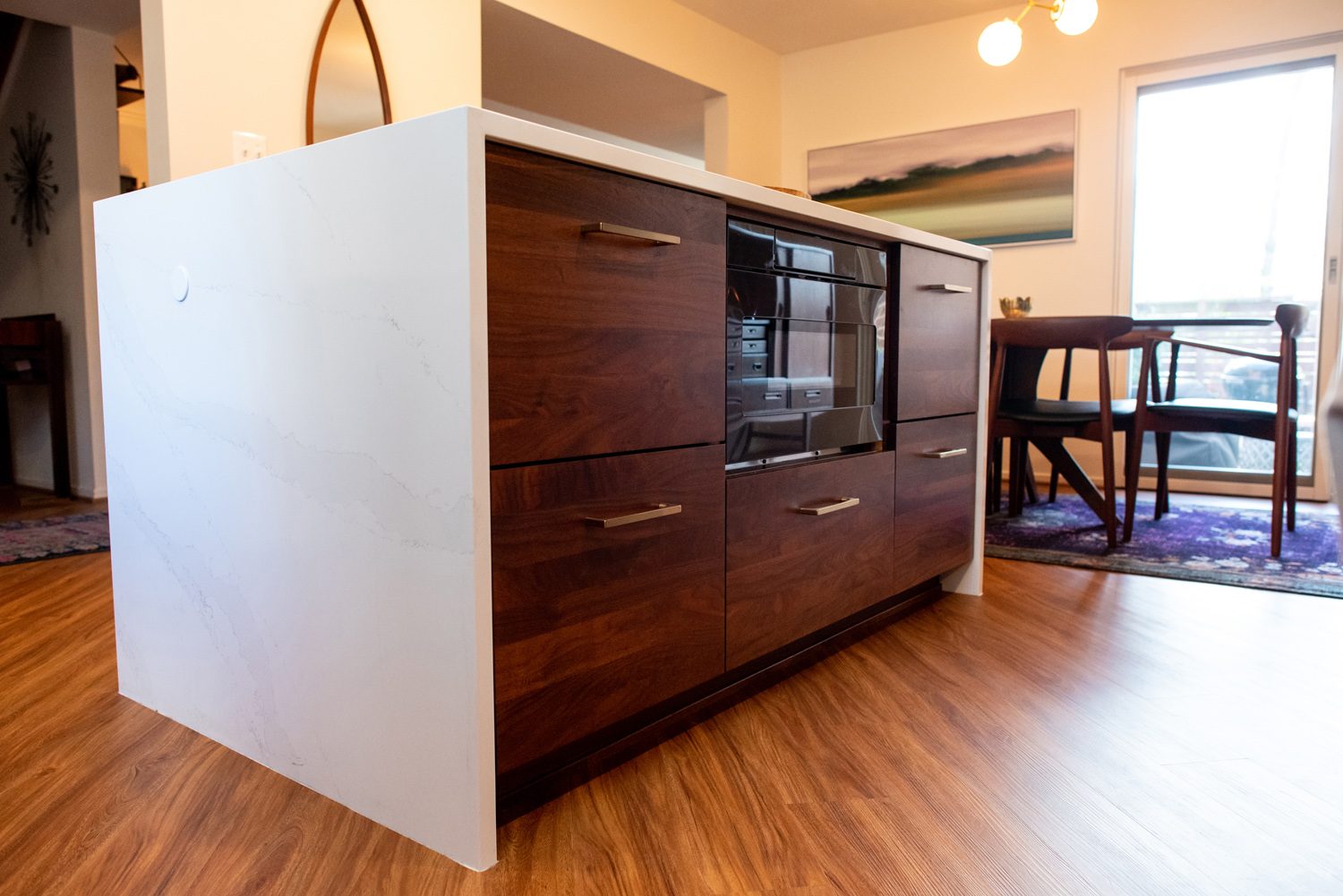 A kitchen island with a white countertop, dark wood drawers, and a built-in oven, situated on wood flooring near a dining area with a table and chairs.