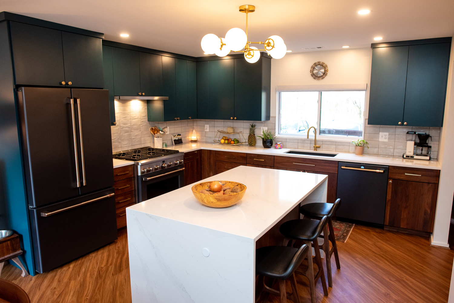 Modern kitchen with dark cabinets, white countertops, a large island with barstools, stainless steel appliances, and wooden flooring, illuminated by ceiling lights.