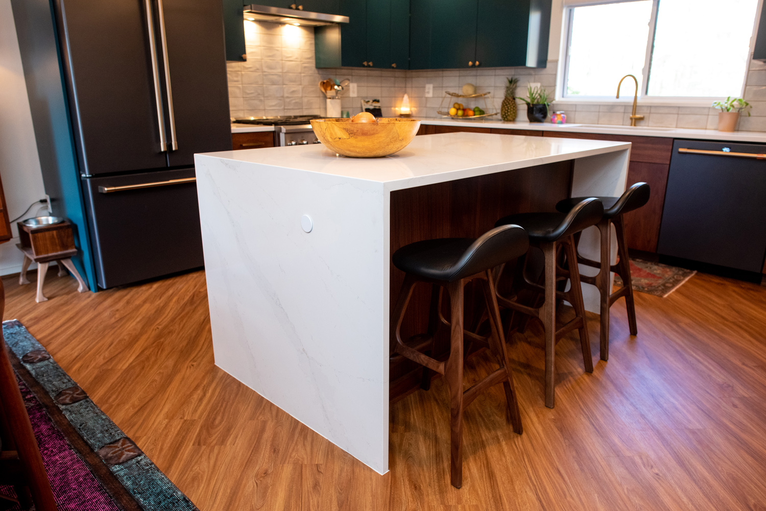 Modern kitchen with a white marble island, three barstools, wood flooring, black appliances, and a fruit bowl on the counter.