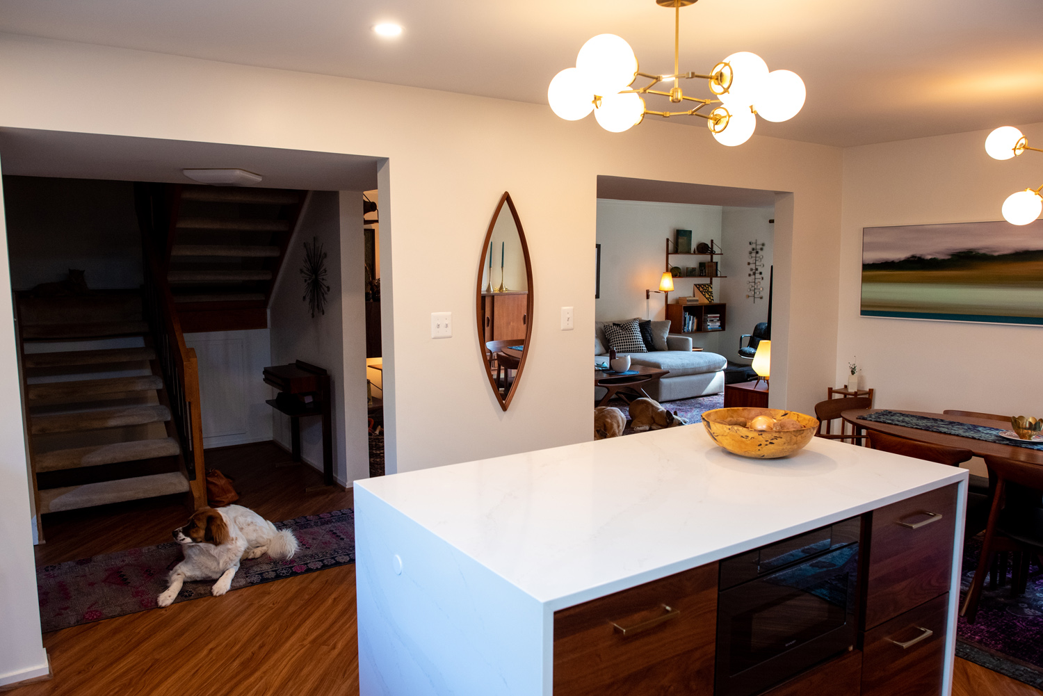 A modern open-concept living area with a kitchen island in the foreground, lounge furniture in the background, and a dog lying on the floor near the staircase.