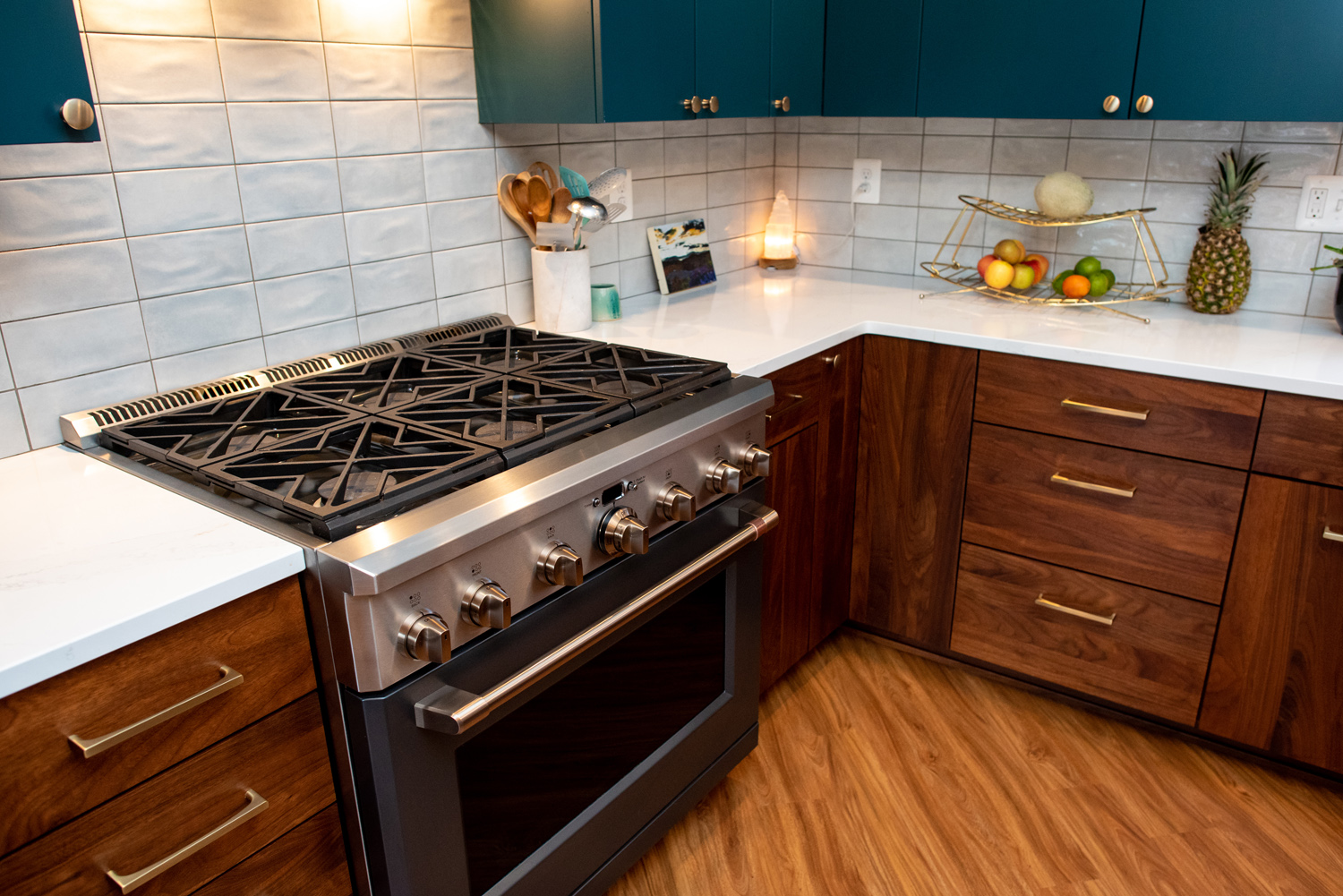 A modern kitchen with a stainless steel gas stove, wooden cabinets, white countertops, teal upper cabinets, and a fruit basket on the counter.
