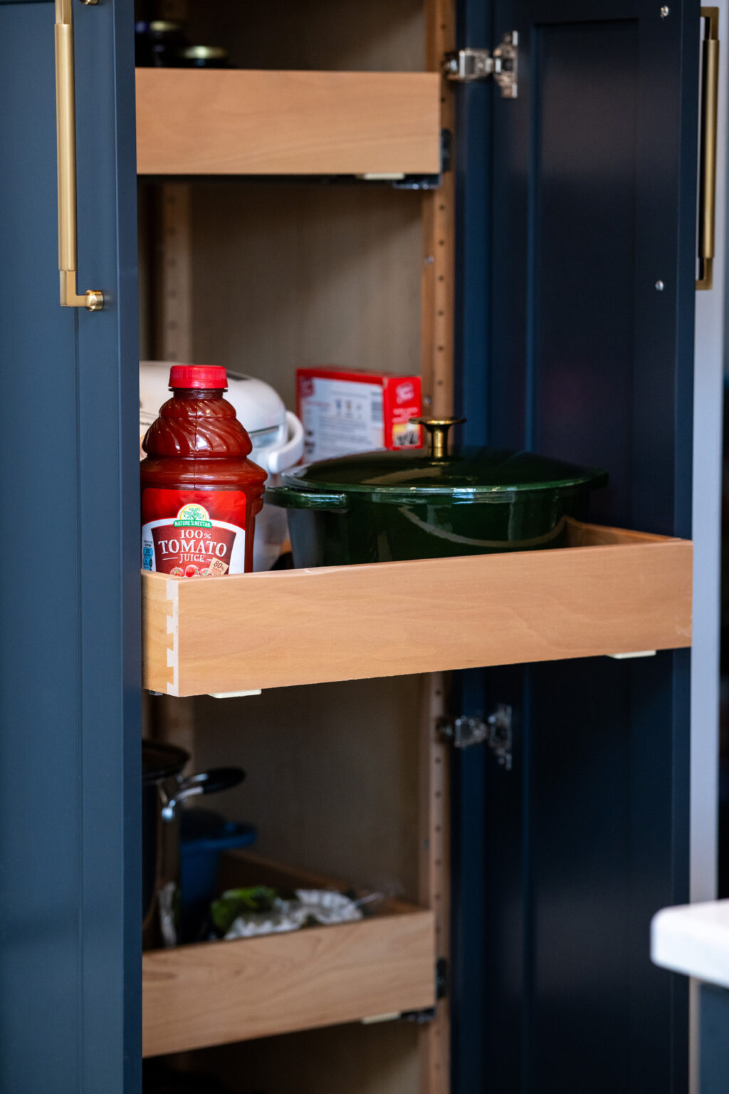 A kitchen cabinet with sliding shelves containing a bottle of tomato sauce, a green pot, and various other items.