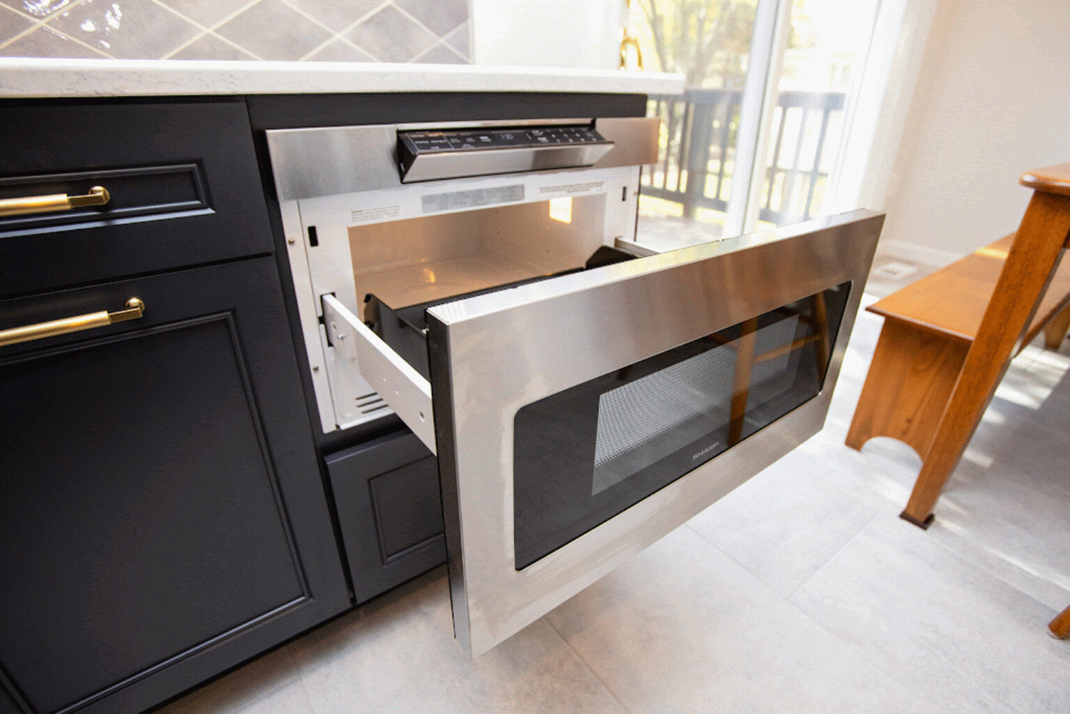 A built-in microwave drawer is partially open in a modern kitchen with black cabinets and a light gray tiled floor.
