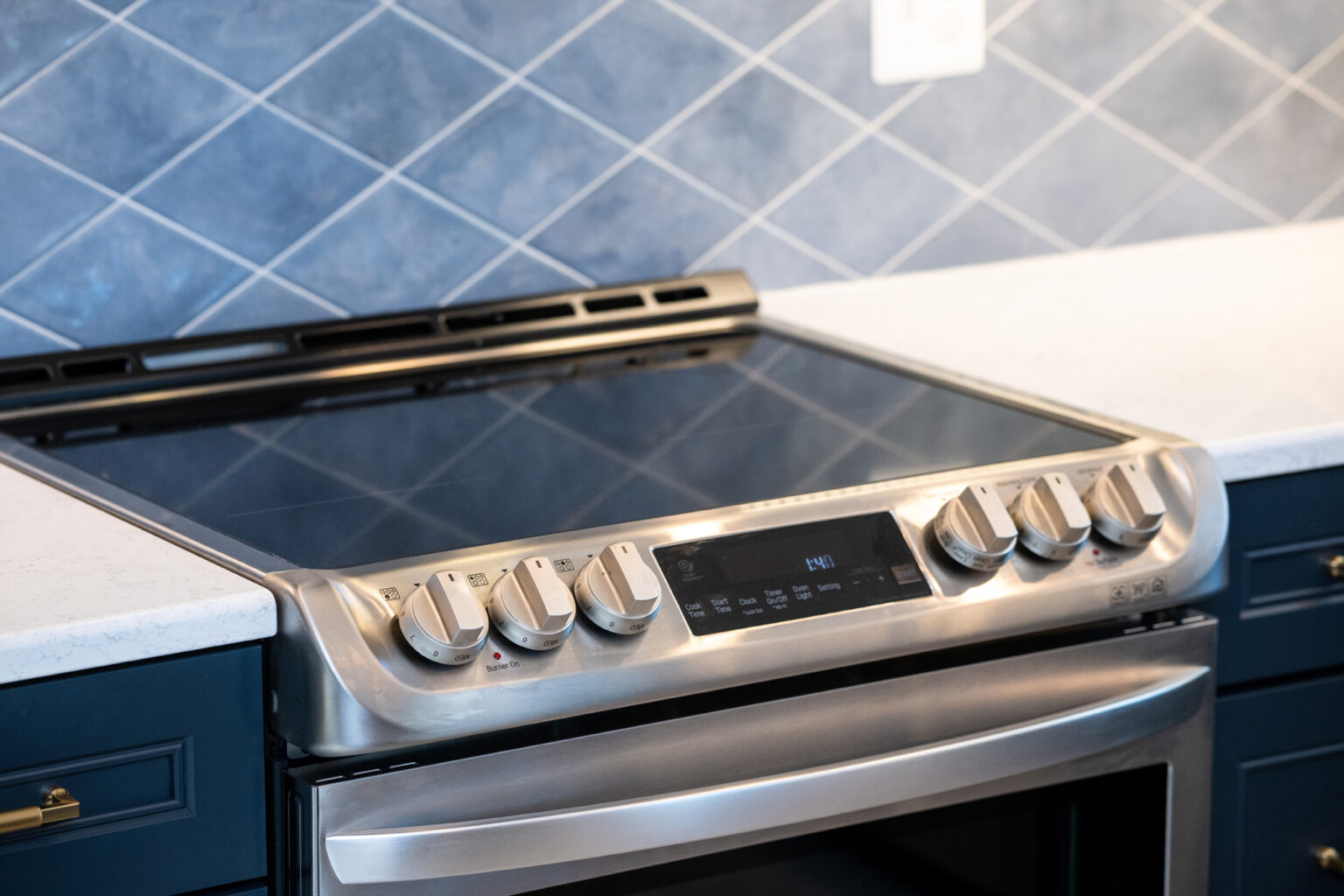 A close-up of a modern stainless steel electric stove with a smooth cooktop and control knobs against a blue tiled backsplash.