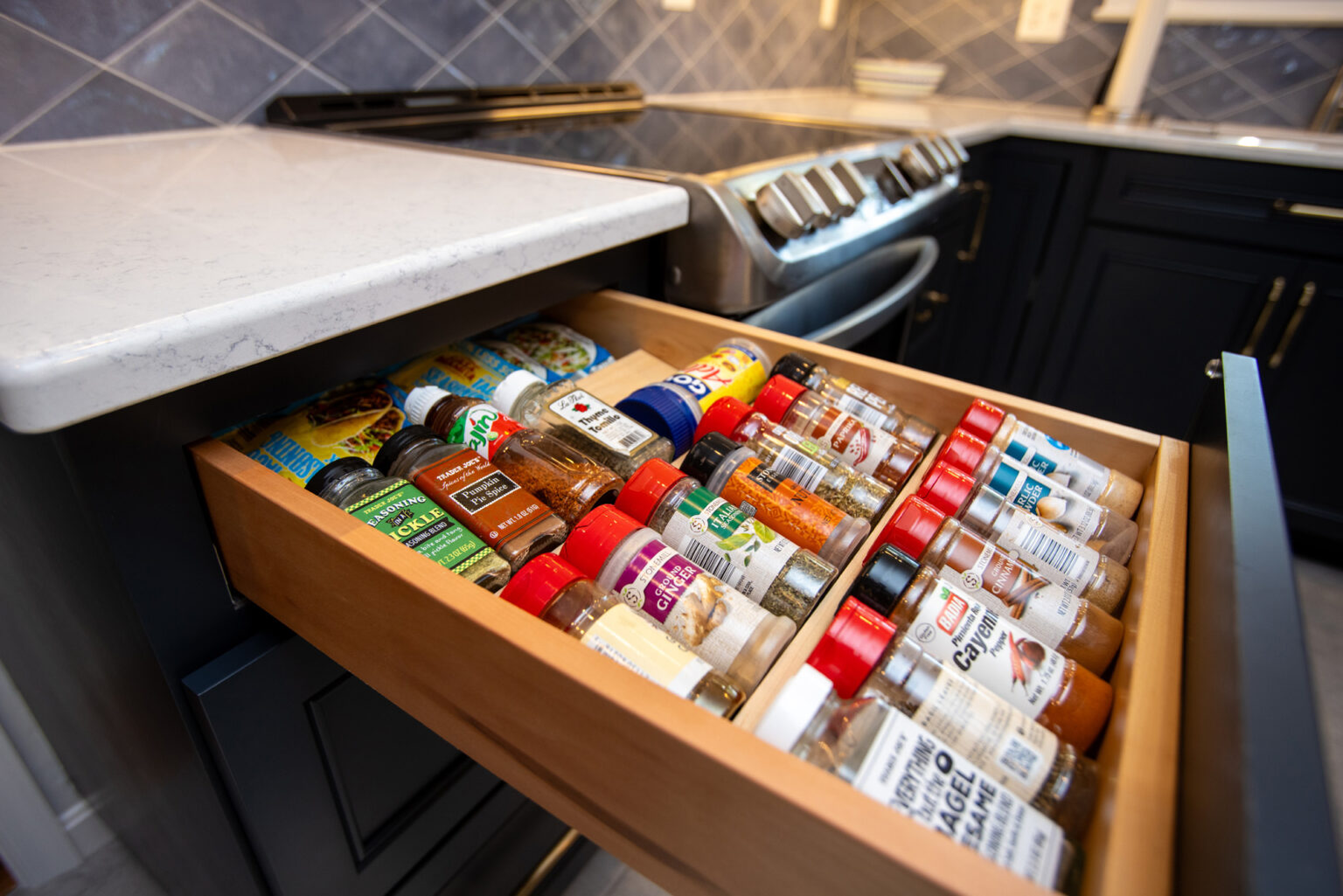 A kitchen drawer is open, revealing an organized assortment of spice jars and containers, placed in a neat row. The background shows part of the kitchen countertop and stove.