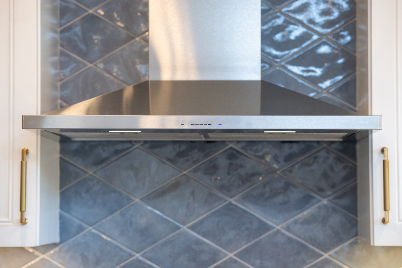 A stainless steel range hood installed above a stove, with blue-gray tiled backsplash and white cabinets featuring gold handles on either side.