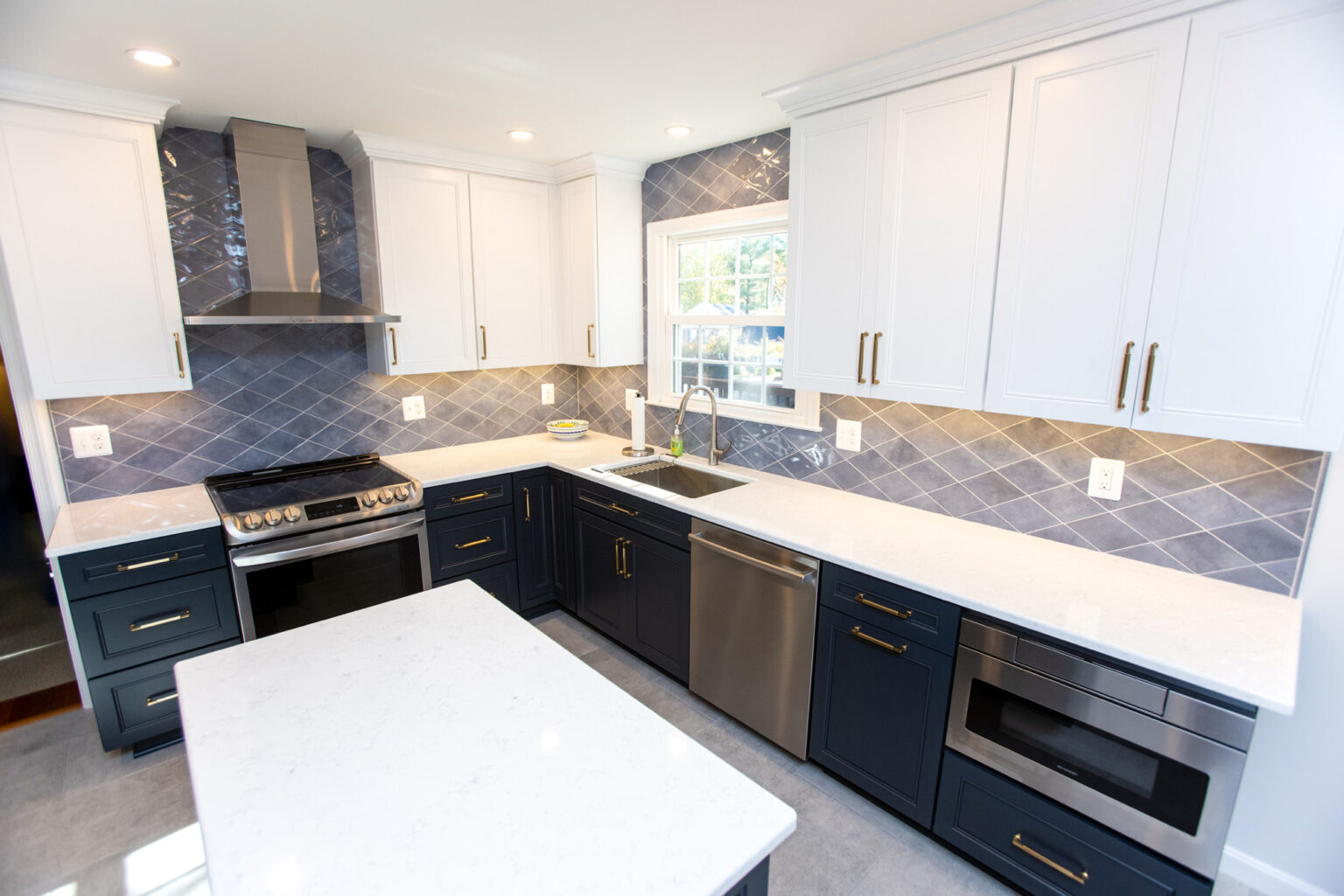 A modern kitchen with white upper cabinets, navy blue lower cabinets, stainless steel appliances, a white countertop, and a blue tile backsplash. A window is above the sink providing natural light.
