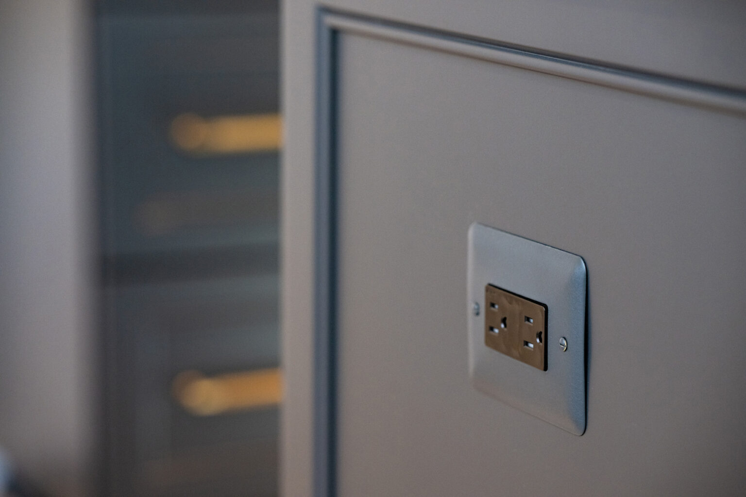 Close-up view of a gray electrical wall outlet with two sockets, set against a smooth, gray wall panel. Blurred golden drawer handles are visible in the background.