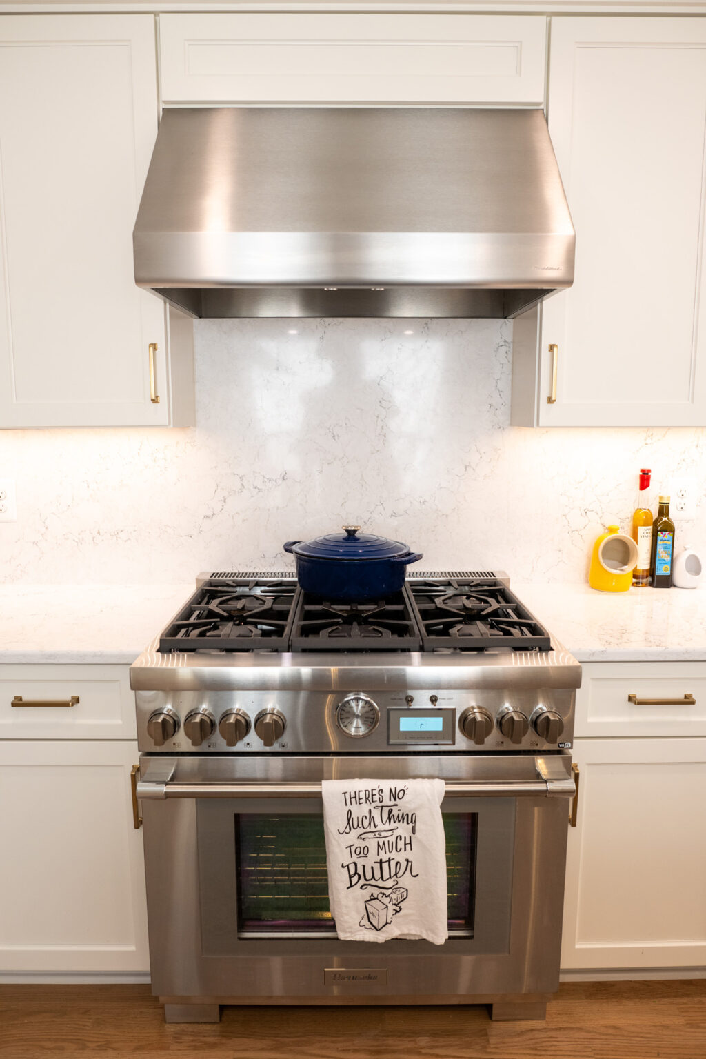 Image by Lesnick Photo A stainless steel gas range with a pot on the stove. A dish towel hanging from the oven handle reads, "There is no such thing as too much butter." The backsplash is white marble.