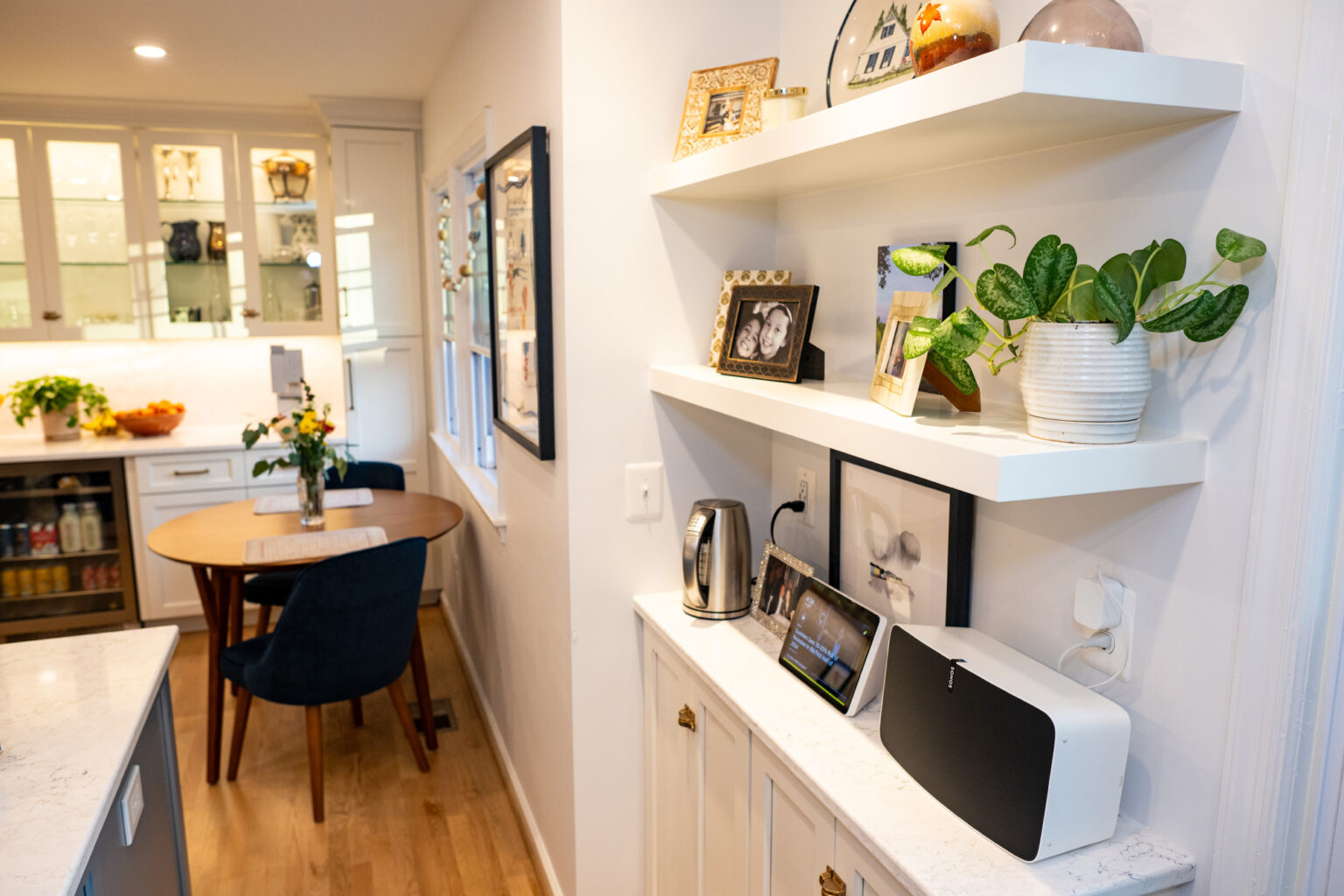 Image by Lesnick Photo A modern kitchen features white cabinets, open shelves with decor and plants, and a round dining table with blue chairs. Appliances are placed on countertops below the shelves.