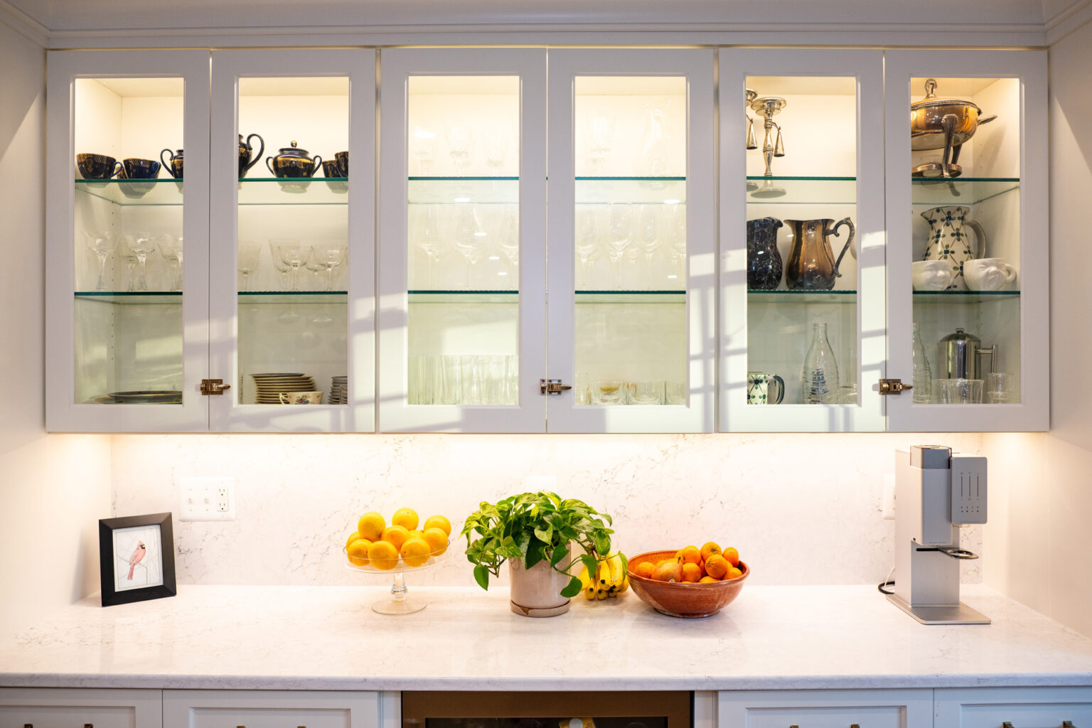 Image by Lesnick Photo A kitchen countertop with bowls of lemons and oranges, potted greenery, framed photo, and a coffee machine. Cabinets above display glassware, teapots, and pitchers.