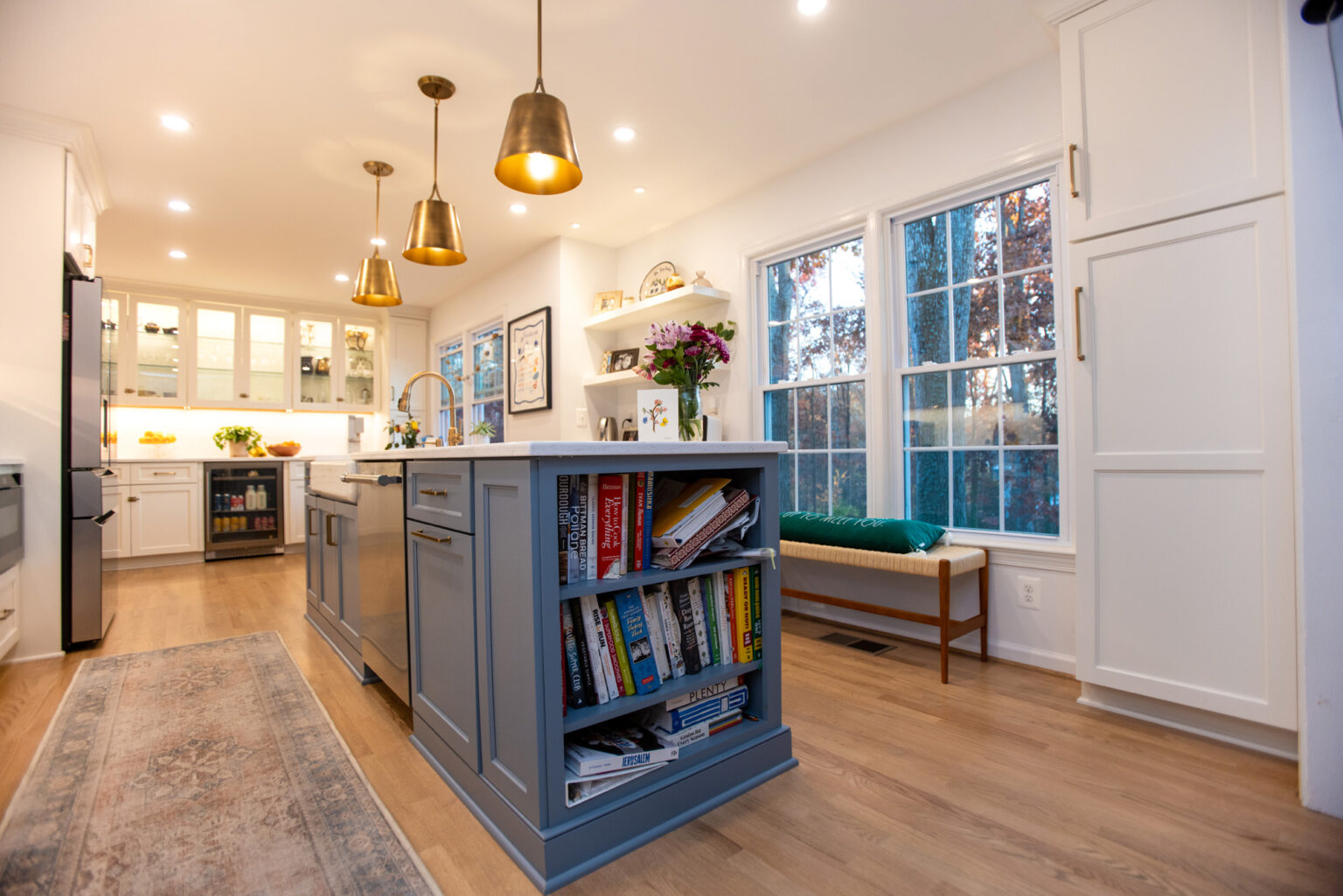 Image by Lesnick Photo A modern kitchen features white cabinets, a large island with built-in bookshelves, pendant lighting, and a window seat with a green cushion. The counter has a vase of flowers and a bowl of lemons.