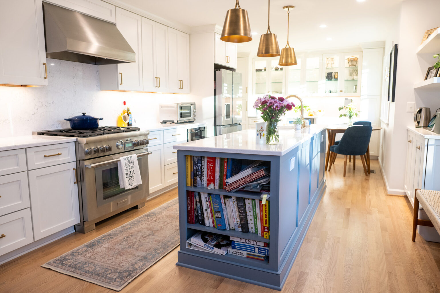 Image by Lesnick Photo A modern kitchen with white cabinetry, stainless steel appliances, a blue island with a bookshelf, and brass pendant lighting. An attached dining area with a table and chairs is visible in the background.