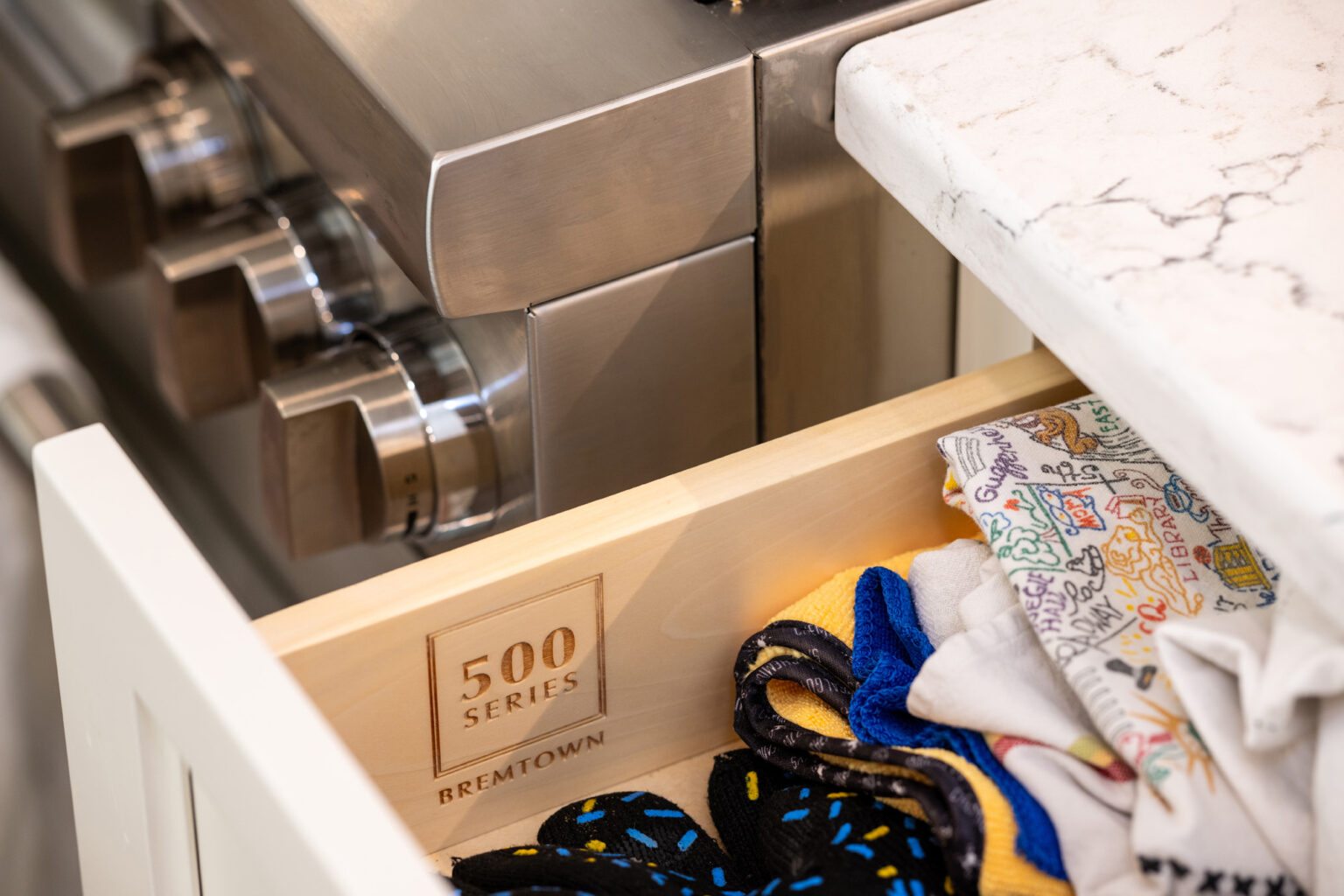 Image by Lesnick Photo A partially open kitchen drawer labeled "500 Series" next to a gas stove. The drawer contains various colorful cloth items.