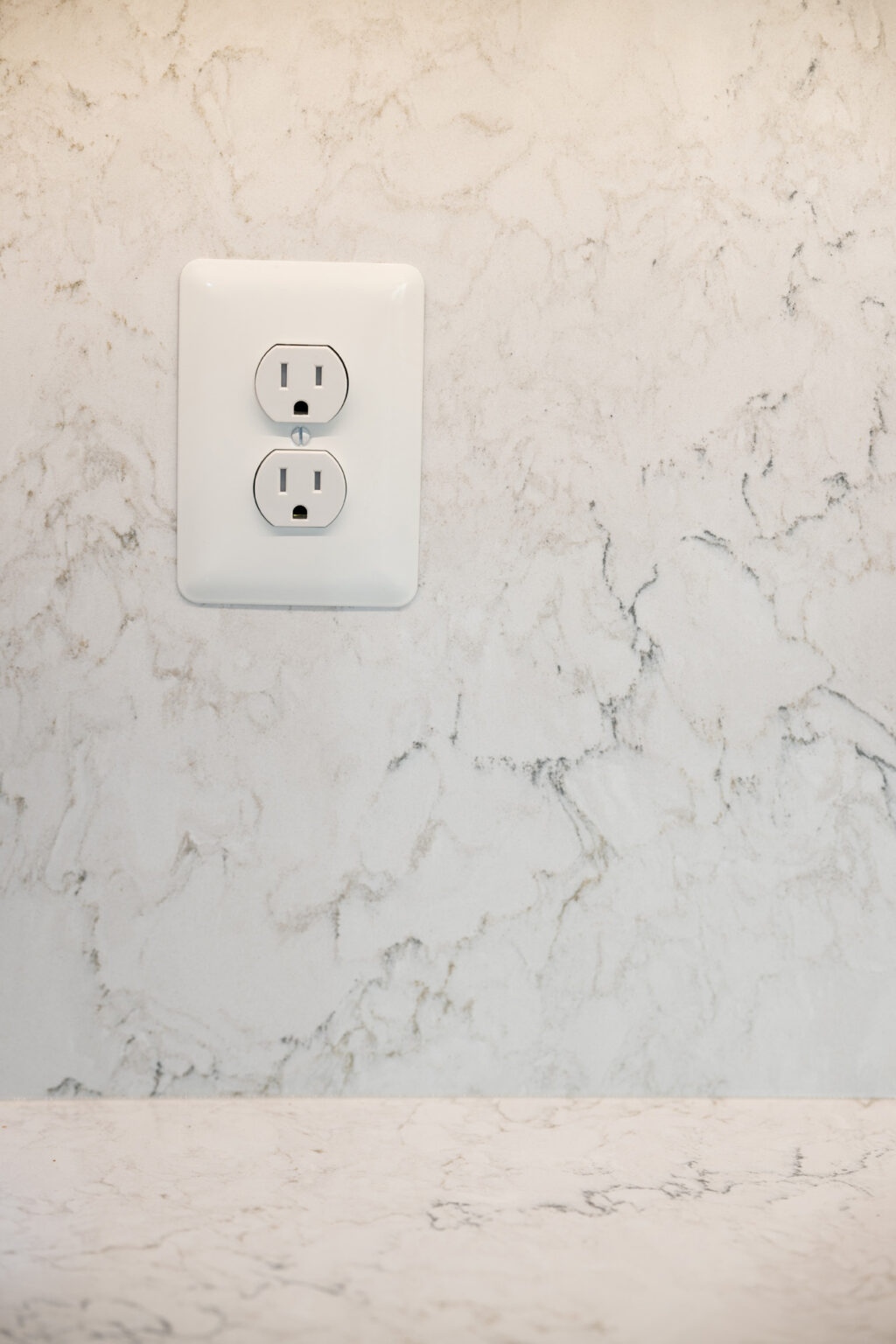 Image by Lesnick Photo A white electrical outlet with two plugs is mounted on a pale, patterned marble wall.
