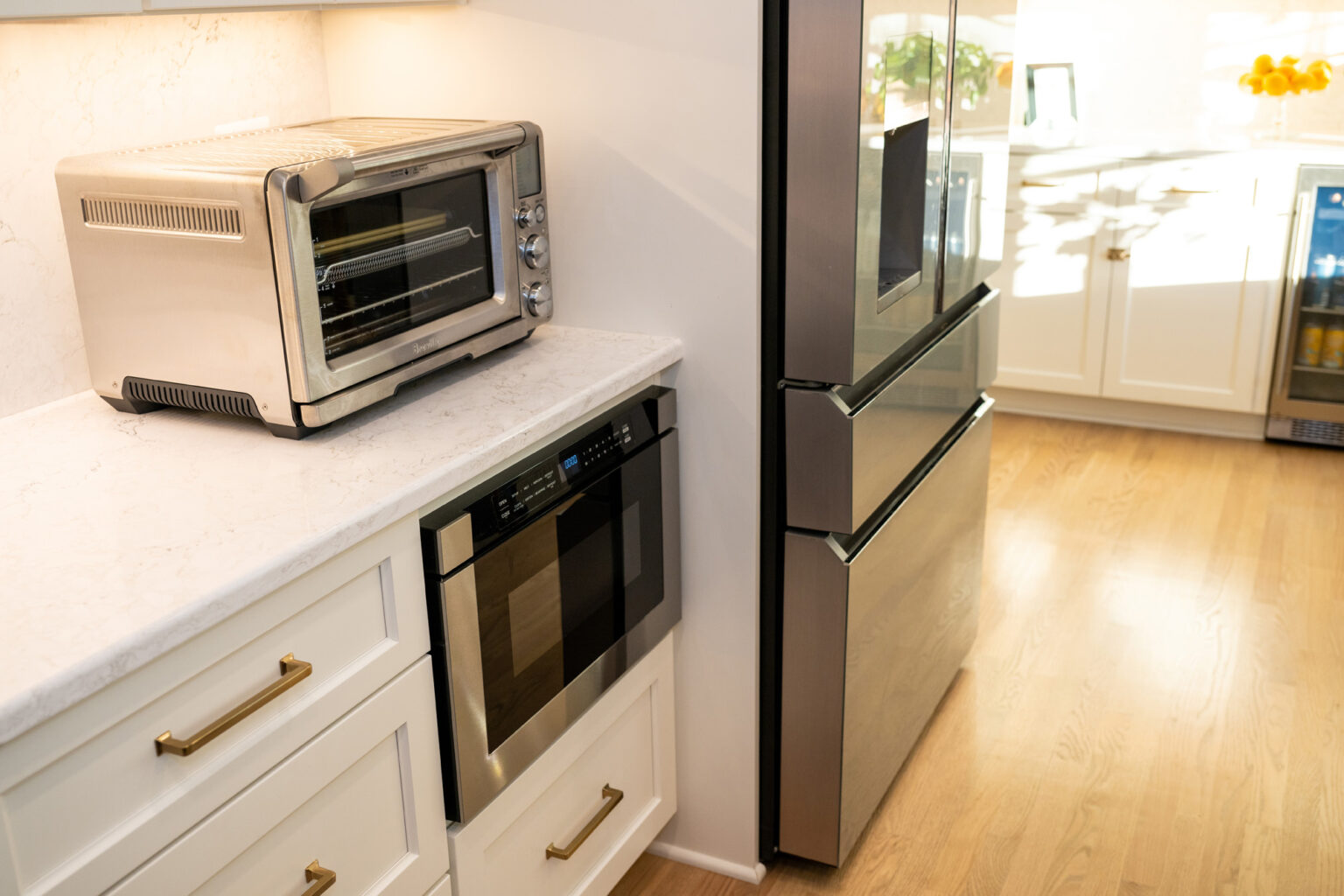 Image by Lesnick Photo A modern kitchen with stainless steel appliances, including a toaster oven, built-in oven, and refrigerator. The countertop is white, and the floor is wooden.