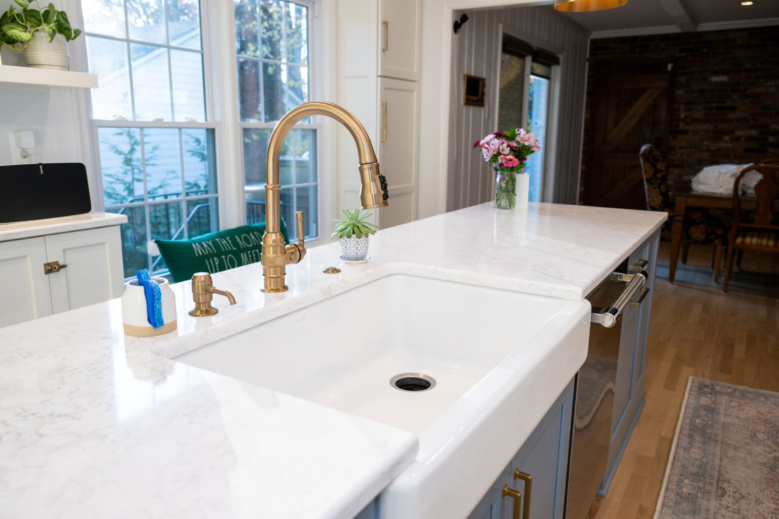 Image by Lesnick Photo A modern kitchen with a large white marble island, a gold faucet, a white sink, and a vase with pink flowers. A window and a barn door are in the background.