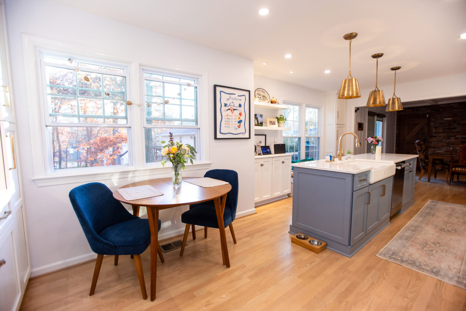 Image by Lesnick Photo A modern kitchen with light wooden floors, a small round table with two blue chairs, a grey island with gold pendant lights, and white cabinets. A vase with flowers and framed art are visible.
