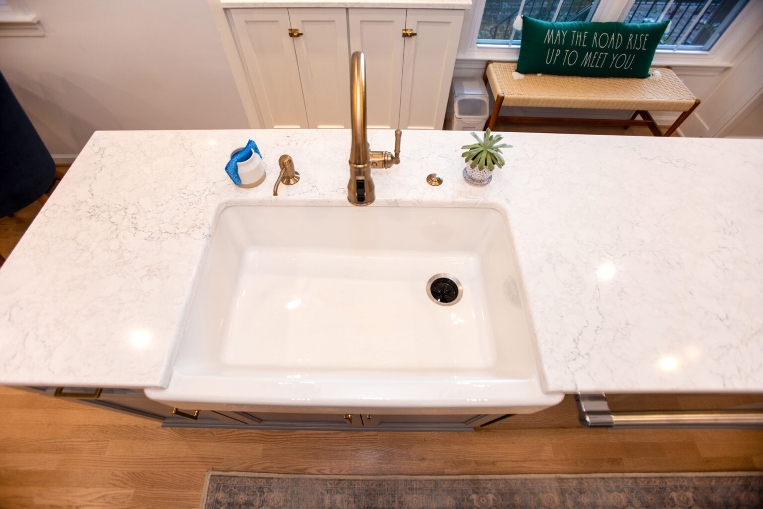 Image by Lesnick Photo A marble countertop with a white sink and gold faucet. A small succulent, soap dispenser, and cleaning sponge are next to the sink. A sign on a bench in the background reads, "MAY THE ROAD RISE UP TO MEET YOU!.