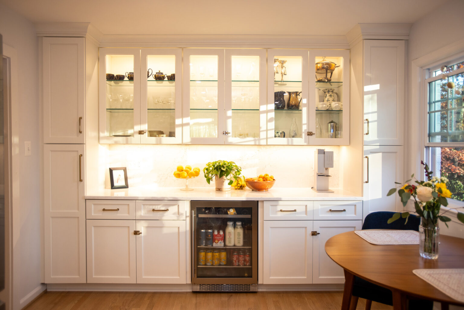 Image by Lesnick Photo A bright kitchen with white cabinetry, glass display cabinets, a small fridge with beverages, a countertop with fruits, a plant, and a framed picture. A window on the right offers natural light.