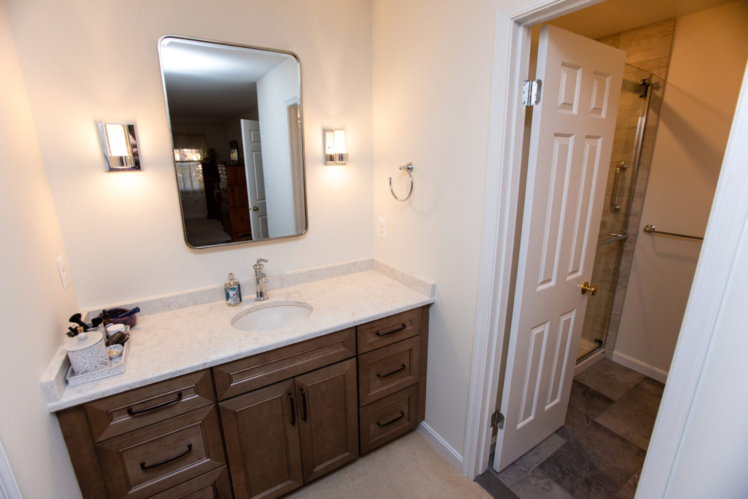 A clean bathroom with a single-sink vanity, mirror, and wall-mounted lights. The door on the right leads to a shower area with beige tiles.